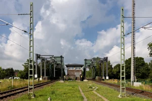 Blick auf zwei parallel verlaufende Bahngleise mit Oberleitungsmasten und einem Stellwerk im Hintergrund unter bewölktem Himmel