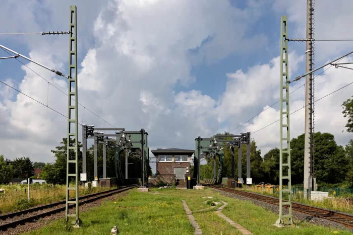 Blick auf zwei parallel verlaufende Bahngleise mit Oberleitungsmasten und einem Stellwerk im Hintergrund unter bewölktem Himmel