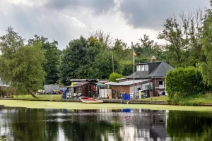 Ufer mit mehreren kleinen Gebäuden, Boot und Bäumen vor bewölktem Himmel, Spiegelung im Wasser