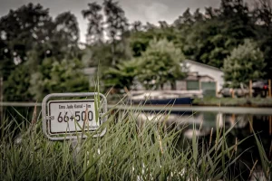 Schild mit der Aufschrift 'Ems Jade Kanal 66+500 km' vor einem Wasserkanal mit Boot und Bäumen im Hintergrund