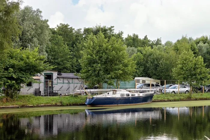 Segelboot am Ufer eines ruhigen Gewässers vor Bäumen, Wohnwagen und parkenden Autos