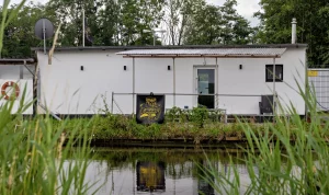 Weißes Haus mit Tür und Fenster steht an einem Wassergraben, davor Gras und Bäume im Hintergrund