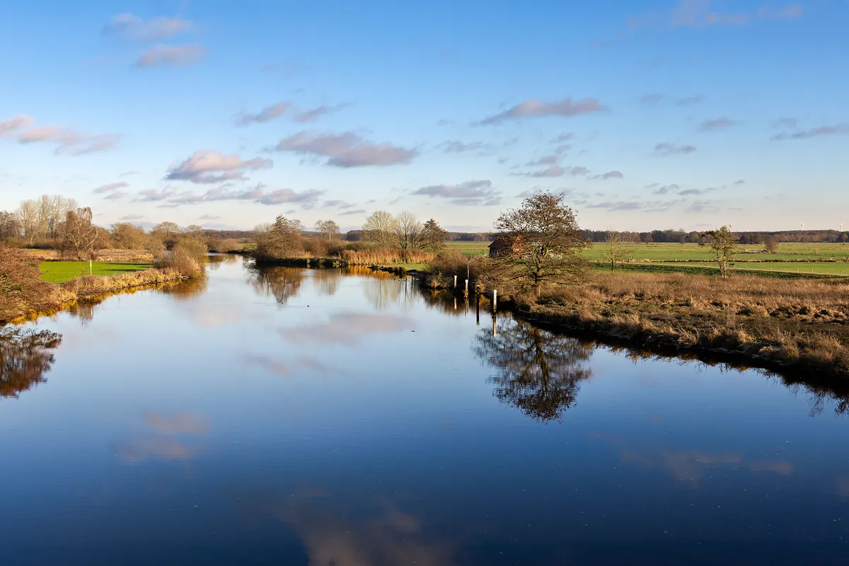 Blick von Tietjens Hütte über die Hamme: ein gewundener Fluss mit grasbewachsenen Ufern und Bäumen unter teilweise bewölktem Himmel, in der Ferne ein kleines rotes Gebäude am Wasser