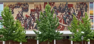 Großes Wandmosaik mit zahlreichen stilisierten Figuren und Symbolen an einer Fassade in Dresden, teilweise von Bäumen verdeckt