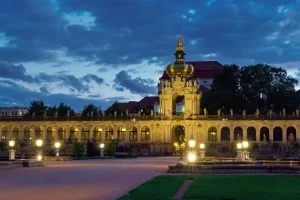 Zwinger-Kronentor abends beleuchtet, Wege und Laternen führen zum Torbogen.