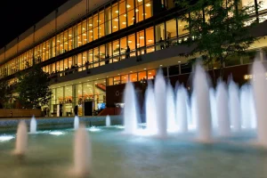 Beleuchtetes modernes Gebäude mit Wasserfontänen im Vordergrund bei Nacht.