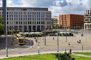 Postplatz mit gelber Straßenbahn, modernen Gebäuden und Fußgängern bei Tageslicht.